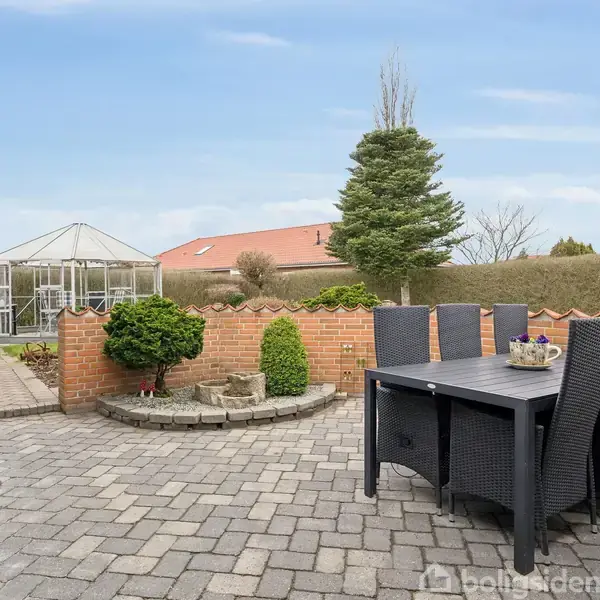Terrasse med sorte havemøbler står på grå stenbelægning. En grøn busk er placeret ved en lav murstensvæg. I baggrunden ses et drivhus og en rød tagbygning.