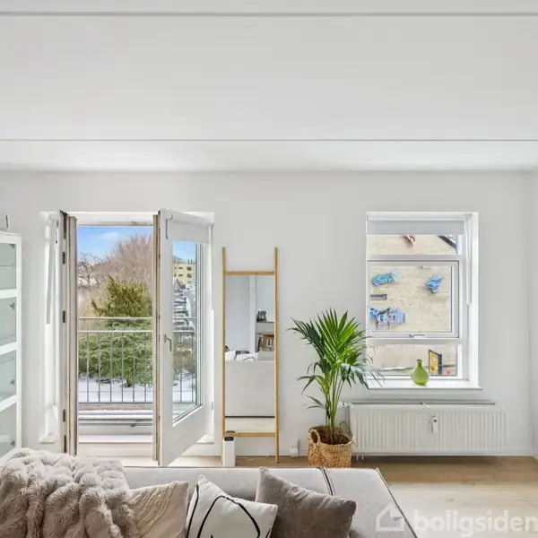 En lys stue med en åben altandør leder ud til en altan. En grønt plante står ved vinduet, omgivet af en sofa og træhylder. Ingen tekst optræder i billedet.