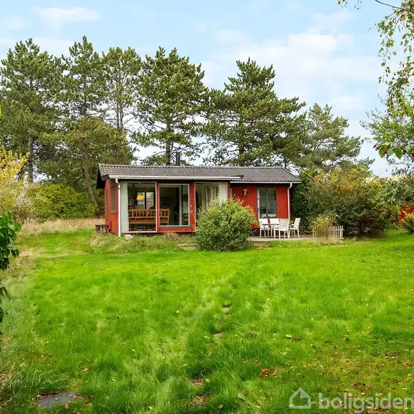 Rødt hus står i en frodig have med grøn plæne og omgivende træer. En terrasse med stole er foran huset, og himlen er blå med lette skyer.