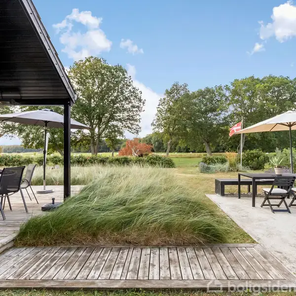 Terrasse med spisebord og stole under en parasol til venstre. Græsbevokset areal i midten, og til højre en siddeplads med parasol. Omgivet af træer og græsmark.