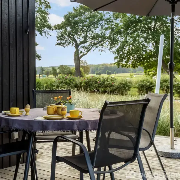 Et udendørs bord står dækket med gule kopper og tallerkener. Bordet er omgivet af sorte stole og beskyttet af en stor parasol, med en mark og træer i baggrunden.