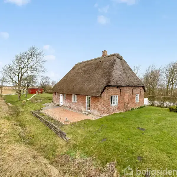 Stråtækt hus står alene på en grøn eng med et terrasselignende område. Omkring ses marker, et træ, og en rød bygning i baggrunden under en lyseblå himmel.