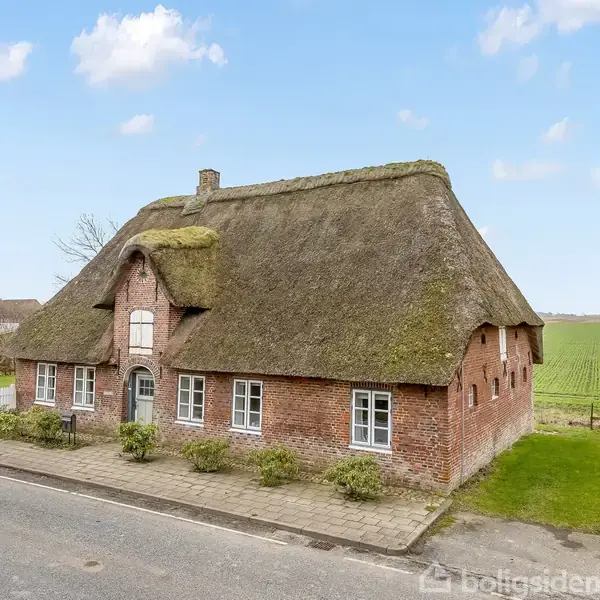 Et stråtækt hus står roligt ved siden af en vej. Det er omgivet af grønne marker og har en rød murstensfacade med flere hvide vinduer.