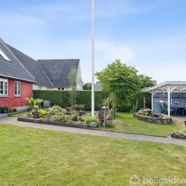 Et rødt hus med hvide vinduer står ved en velplejet have med grøn græsplæne. Til højre en terrasse med et shelter og drivhus omgivet af planter.