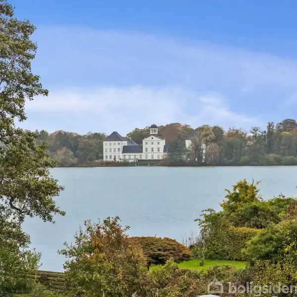 Et hvidt slot står ved søbredden, omgivet af træer og grøn natur. Blå himmel i baggrunden.