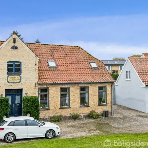 Et gult murstenshus med rødt tegltag står ved siden af et hvidt anneks. En hvid bil er parkeret foran. Et skilt på facaden siger "Købmand". Græsområde i forgrunden.