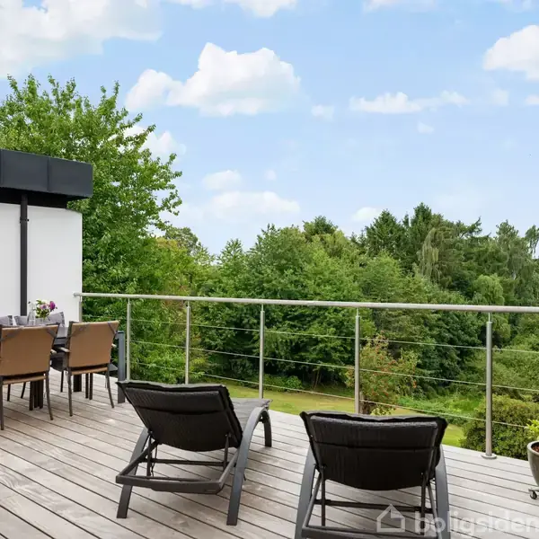 To liggestole og et spisebord med stole står på en træterrasse. Terrassen har glasrækværk og udsigt til grønne træer under en blå himmel med få skyer.
