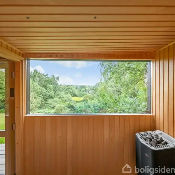 Sauna med træbeklædning står åben mod en stor vinduesudsigt, der viser frodig grøn natur med træer og græsplæner. En varmeovn er placeret til højre i rummet.