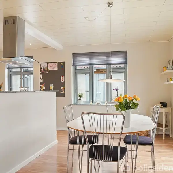 Et spisebord med fire stole står i et lyst køkken-alrum med trælaminatgulv. En bordlampe hænger over bordet, og blomsterdekorationer pynter vindueskarme og bord.