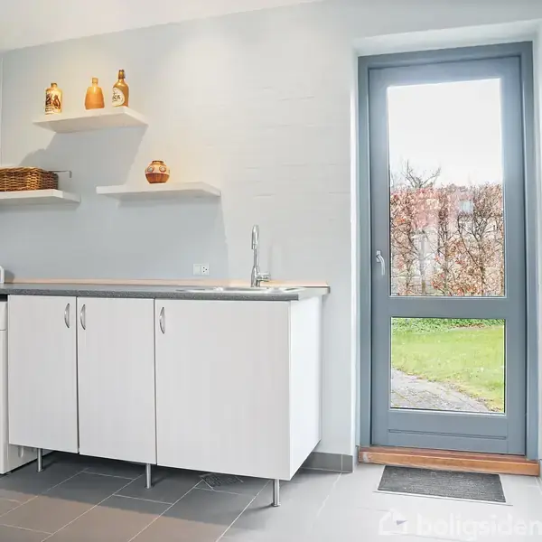 En vaskemaskine står ved siden af hvide skabe under en bordplade. Over bordpladen hænger hylder med krukker og en kurv. En glasdør fører ud til en græsplæne.