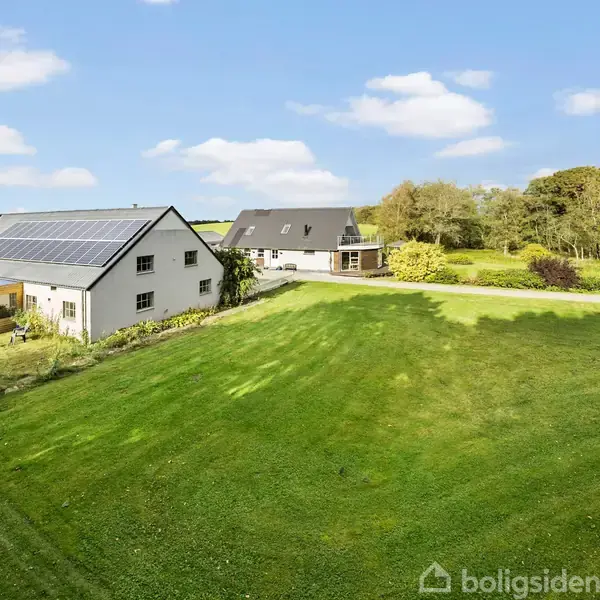 Et hvidt hus med solpaneler på taget står på en stor grøn plæne. I baggrunden er der træer og en blå himmel med få skyer.