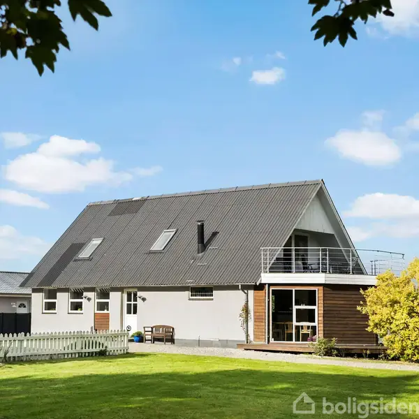Et moderne hus med skråt tag og flere tagvinduer står i en velplejet have. Ved siden af huset er en terrasse med gelænder. Himlen er blå med spredte skyer.
