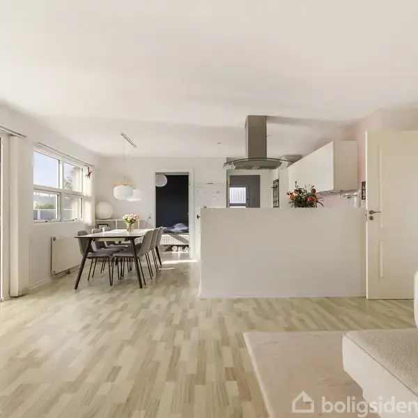 Stue med sofa i forgrunden, spisebord og stole i midten, køkken bagved. Store vinduer og glasdøre giver udsigt til terrasse med planter, fyldt med naturligt lys.
