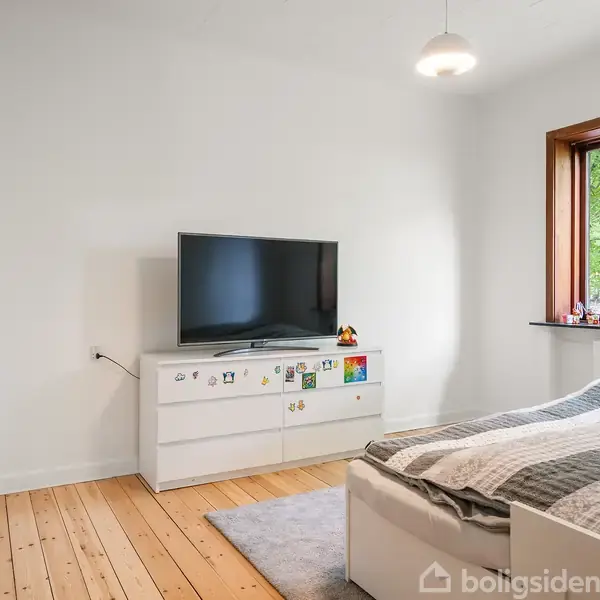 En hvid kommode med et tv ovenpå står i et lyst soveværelse. Der er en seng med gråt sengetøj ved siden af, og et vindue viser grønne træer udenfor.