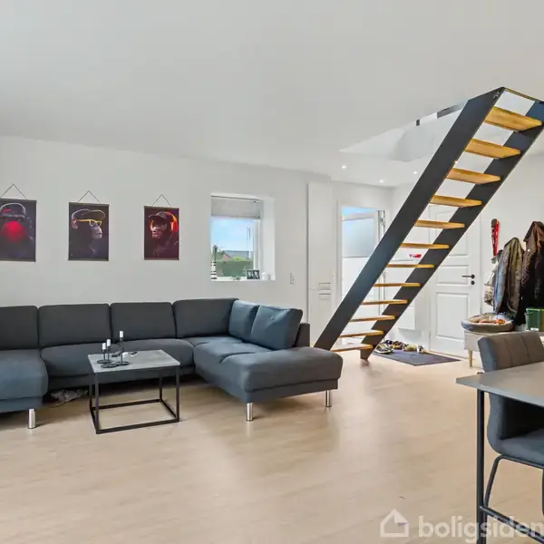 En grå hjørnesofa står i en lys stue. Tre billeder hænger på væggen. En trappe fører til en anden etage. Ved siden af står en spisebordstol.