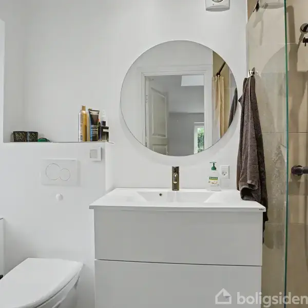 Badeværelse med hvid vask og skab under rundt spejl. Bruser i glasafskærmning til højre. Toilet ved siden af vindue til venstre. Håndklæde og sæbedispenser på vask.
