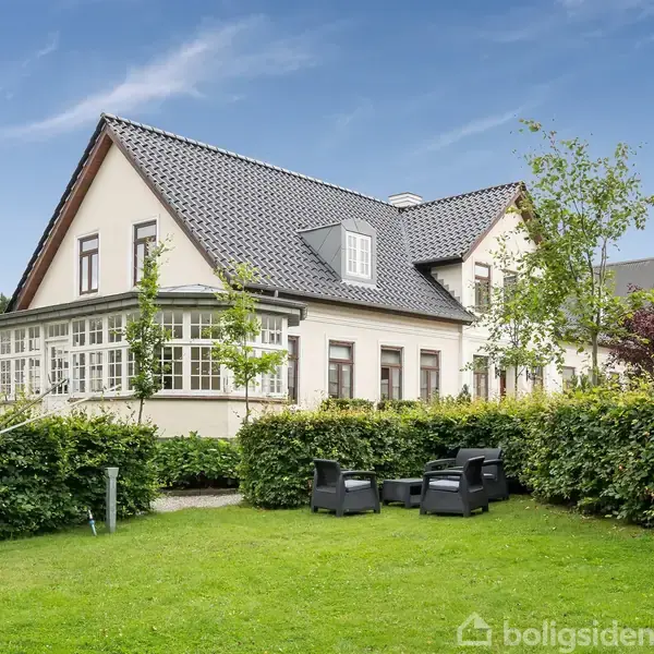 Et hvidt hus med mørkt tag står i en velplejet have. Tre sorte havestole er placeret på græsplænen omgivet af grønne hække og træer under en blå himmel.