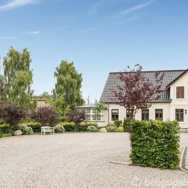 Hvid villa med sort tag står i en gårdsplads omgivet af grus og grønne buske. Til venstre ses en flagstang med dansk flag, omgivet af træer.