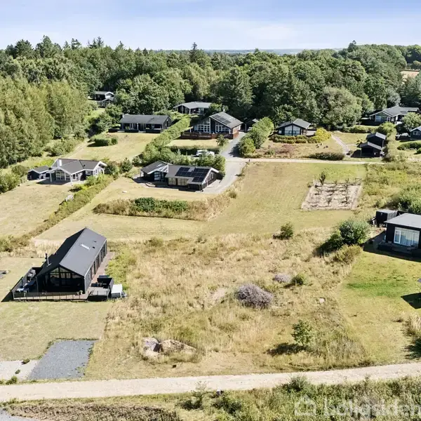 Luftfoto viser en række huse placeret tæt på en stor sø omgivet af grønne marker og skove. Bagsiden af en ejendom er afgrænset, mens søen strækker sig i baggrunden.