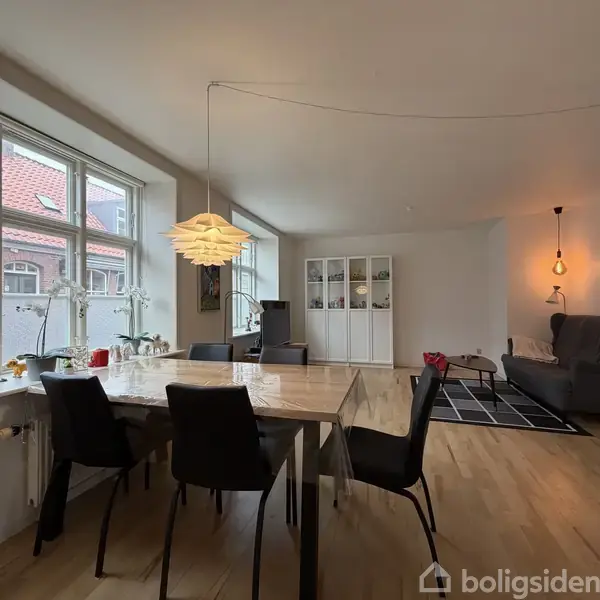 Et spisebord med sorte stole står i et lyst rum med brændeovn og sofaområde. Store vinduer giver udsigt til en bygning med rødt tag. En lampe hænger over bordet.