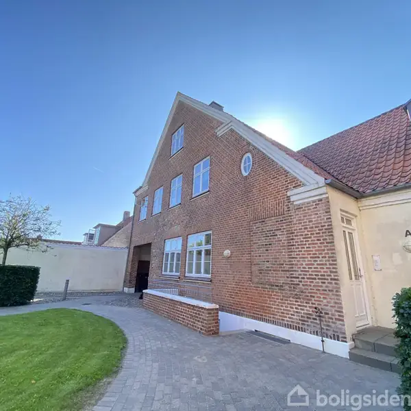 En stor murstensbygning med skråt tag står i solrigt vejr. Omkranset af en brostensbelagt sti og en græsplæne med en enkelt træ og hæk i baggrunden.