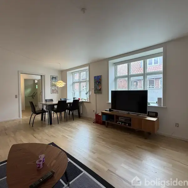TV står på et moderne træbord ved siden af store vinduer i et lyst, rummeligt opholdsrum. Et spisebord med stole ses i nærheden, omgivet af hvide vægge og trægulv.