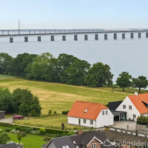 Huse med røde tage står stille i forgrunden ved en grøn mark. I baggrunden ses en lang bro over en bred vandstrækning med træer langs kysten.