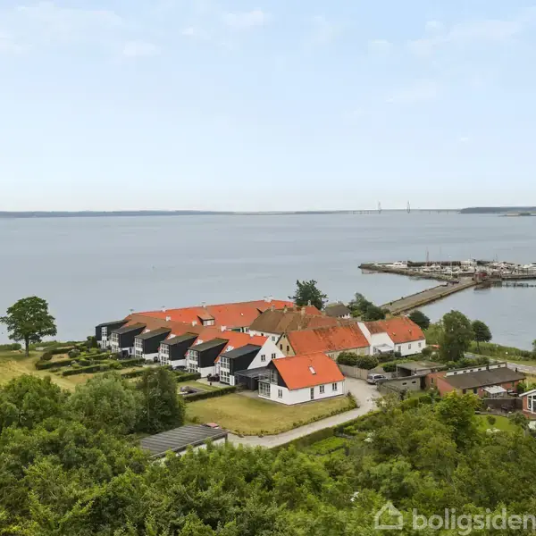Rækkehuse med orange tage står stille ved kysten, omgivet af grønne områder og en mole, der strækker sig ud i vandet. En bro kan ses i det fjerne.