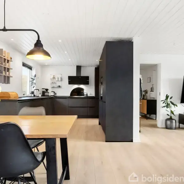 Spisebord med sorte stole står i køkken-alrum under sorte lamper. Køkkenet har mørke skabe og industrielt design. Ved siden af ses en stue med en stor fladskærms-TV.