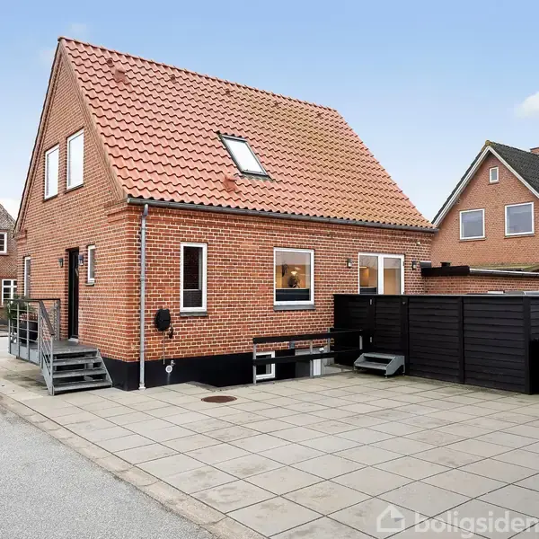 En rød murstenshus med tagvindue, trappe ved siden, placeret langs en stille gade med flisebelagt fortov og skraldespande til højre. Vejskilt i baggrunden.