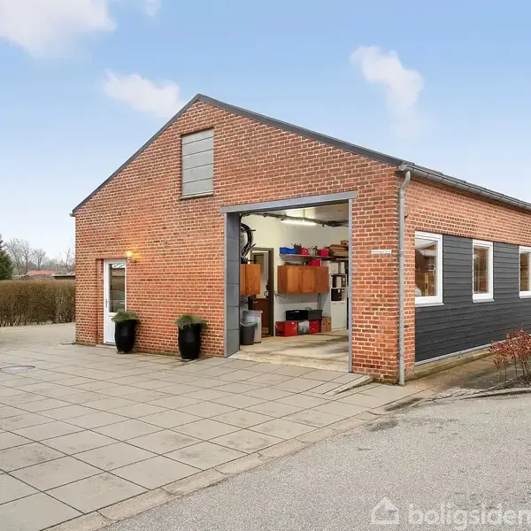 Rød murstensbygning med åbent garageport, hvor forskellige værktøjer og genstande er synlige. Bygningen er omgivet af flisebelagt område og en parkeringsplads med en sort bil i baggrunden.