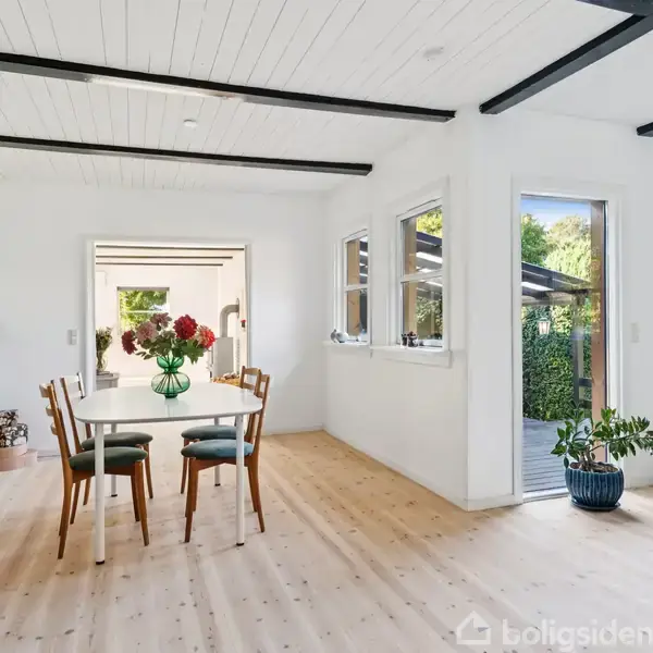 Spisebord med fire stole står i et lyst rum. Bordet er pyntet med røde blomster. Store vinduer og en åben dør viser udsigt til en terrasse med planter udenfor.