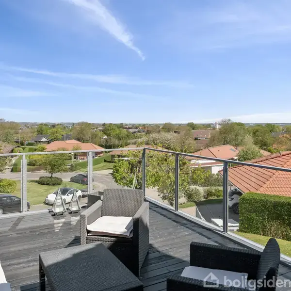 Terrassemøbler står på en balkon med glasrækværk. De viser en rolig aktivitet i et boligområde med udsigt over huse og grønne træer under en klar blå himmel.