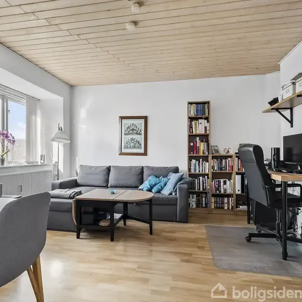 En stue med en grå sofa og sofabord, placeret foran en reol. Et skrivebord med computer står ved siden af. Rummet har lyse trægulve og et vindue med udsigt.