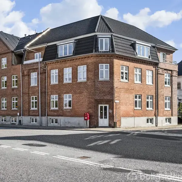 En rød murstensbygning med sort tag står i gadehjørnet. Bygningen har flere vinduer og en hoveddør. Omgivet af en vej og blå himmel med skyer.