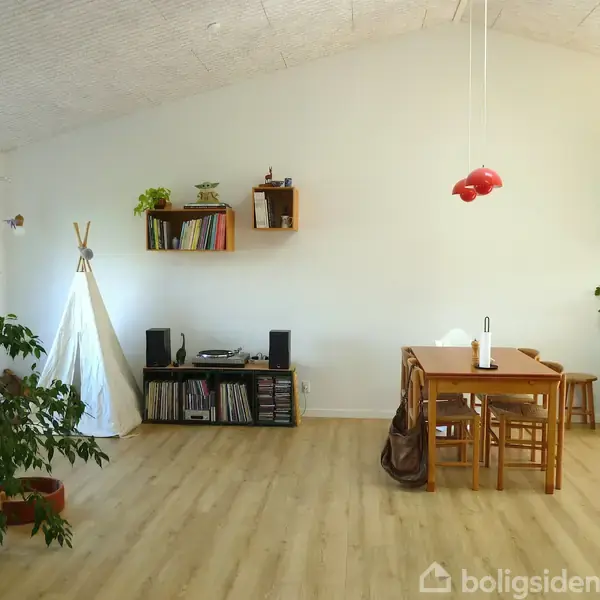 Et lyst rum med trægulv indeholder et spisebord med stole, en tipi, og en reol med bøger og planter. En pæne hængelampe og en plante i krukke er til stede.