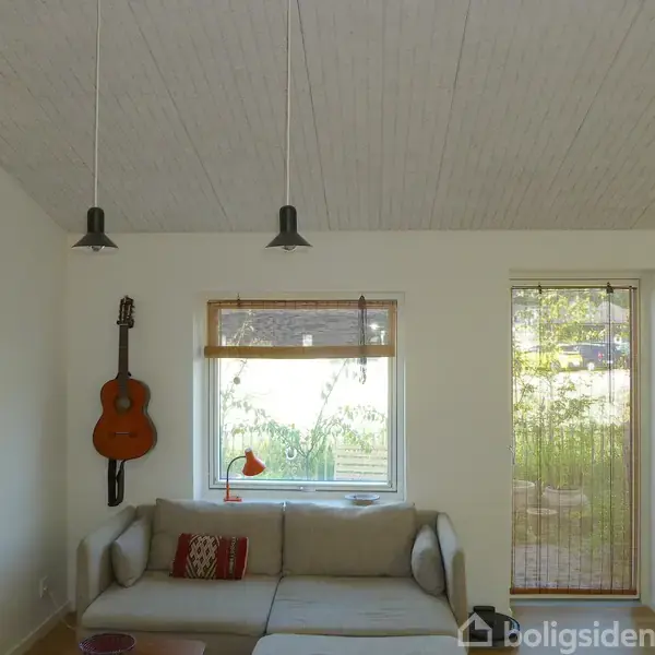 En beige sofa står under et vindue med en orange lampe i en lys stue. En guitar hænger på væggen ved siden af et farverigt maleri.