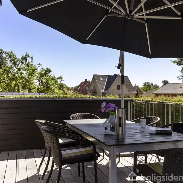 En terrassebord med seks stole står under en stor, sort parasol. En vase med blomster og nogle mapper pryder bordet. Omgivet af grønne træer og hustage i baggrunden.