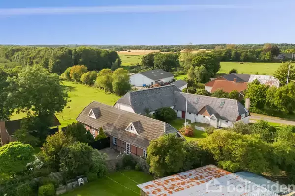 Lystejendom omgivet af stor have og marker. Hovedbygning med rødt murværk og gavlvinduer. Flere udhuse bagved og frodig natur i baggrunden.