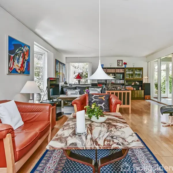 Stue med rød lædersofa og marmorbord, hvorpå står et lys og en plante. Omgivet af store vinduer med udsigt til grønne planter, kunstværk på væggen, og reol i baggrunden.