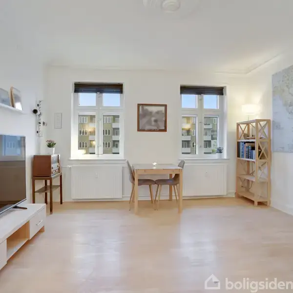 TV på reol står i stue med trægulv. Spisebord med stole opstillet foran to vinduer. Hylde med billeder hænger på væg. Reol med bøger og kort til højre.