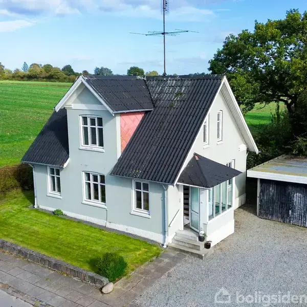 Et hvidt hus med sort tag står på en velplejet græsplæne. Der er en enkelt garage til højre. Baggrunden viser åbne marker og nogle træer.