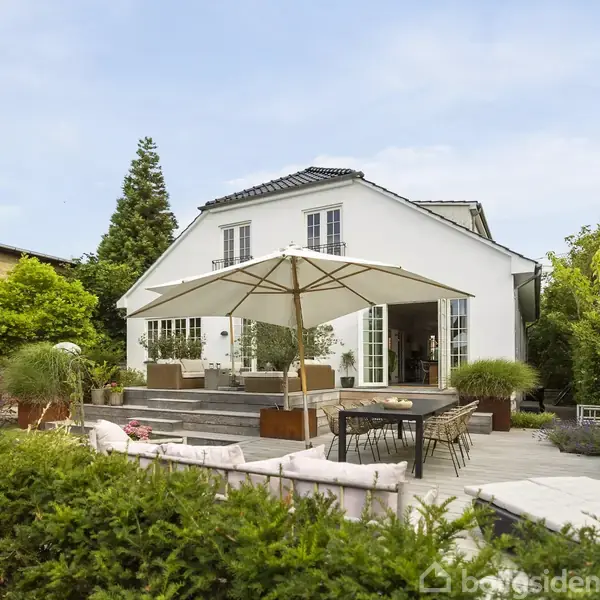 Hvidt hus med fransk altan har terrasse med udendørs sofaer og parasol. Omgivet af have og planter, står spisebord med stole på trædæk. Skyfri himmel i baggrunden.