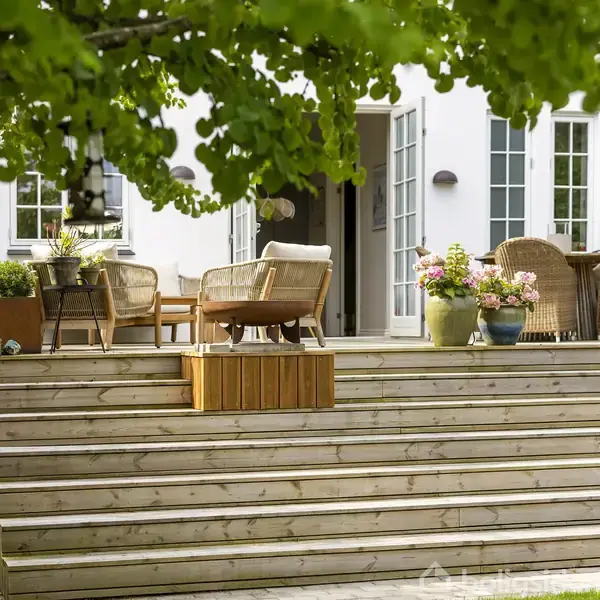 En terrasse med træmøbler og blomsterkrukker står ved en hvid bygning med åbne franske døre. Grønne træer omkranser området og skaber en hyggelig atmosfære.