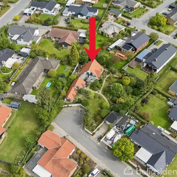 Et rødt pil peger ned på et hus med en rød tag i en boligområde med flere huse. Omkringliggende græsplæner og træer er synlige langs vejene.