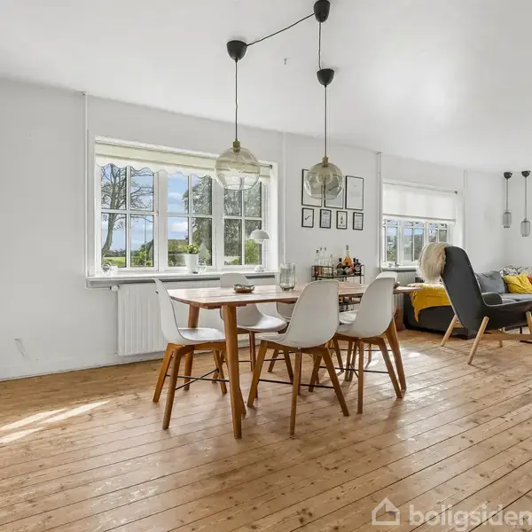 Spisebord står i stuen med stole omkring. Store vinduer lader sollys ind, oplyser rummet med trægulv og reoler. Lampetter hænger over bordet og ved sofaen.