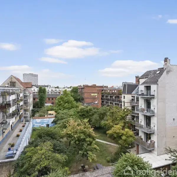 Flere etagebygninger omkranser en grøn gårdhave med træer og buske, hvor altaner strækker sig ud fra bygningerne. Blå himmel med skyer udgør baggrunden i bymiljøet.