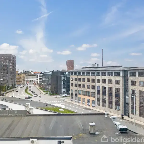Bygninger står stille langs en vej i en moderne by. Det er klart vejr med spredte skyer på himlen.