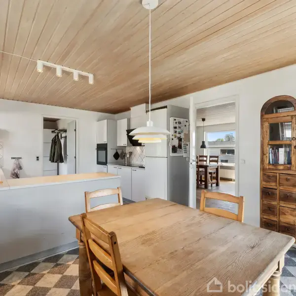 Træbord med fire stole står under en hængende lampe i et køkken med lyst træloft. Skabset over bordet; åbne åbninger fører til andre rum. Ternede gulvfliser.