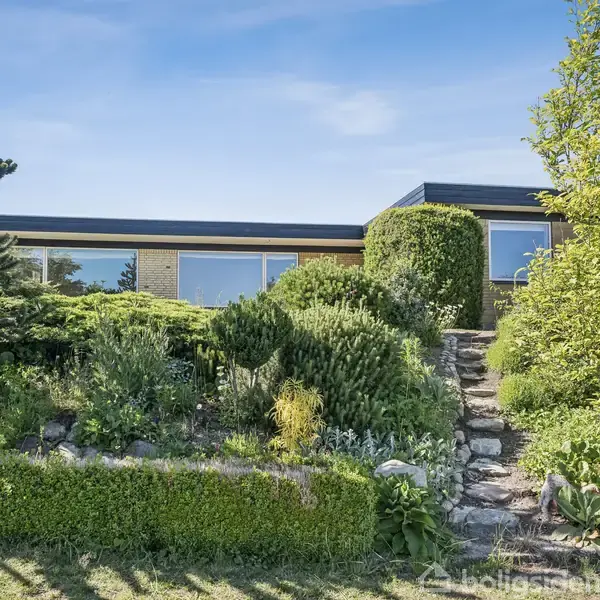 Et moderne hus ligger bag en frodig have med forskellige buske og træer. En stensti fører gennem det grønne til hovedindgangen. Blå himmel ses ovenover.